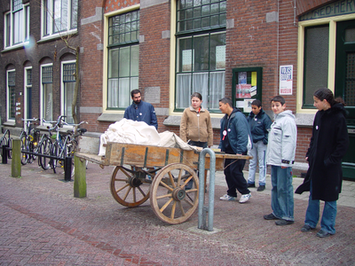 879486 Afbeelding van enkele leerlingen van een middelbare school die proberen een handkar aan te duwen, voor het ...